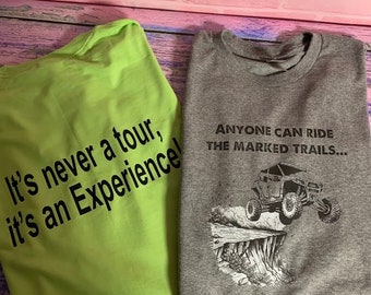 May include: Two t-shirts with different designs. The green t-shirt says "It's never a tour, it's an Experience!" The grey t-shirt has a black and white illustration of an off-road vehicle driving off a cliff and says "Anyone can ride the marked trails... Only a few can ride with the Mudman."