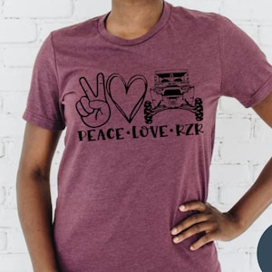 May include: A maroon t-shirt with a peace sign, heart, and a black and white illustration of an off-road vehicle. The text "Peace Love RZR" is printed below the graphic.