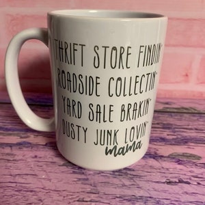 May include: White ceramic mug with black text that reads "Thrift Store Findin', Roadside Collectin', Yard Sale Brakin', Dusty Junk Lovin' mama". The mug has a curved handle and is set against a pink and purple background.