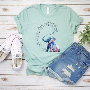 Op de afbeelding: Een tealblauw t-shirt met een aquarel olifant ontwerp en de tekst "Live without fear, Love without limits".
