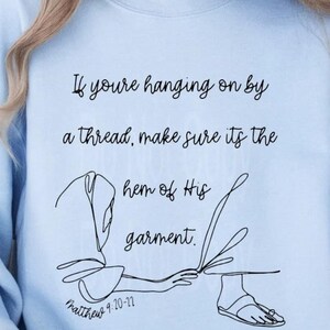 Op de afbeelding: Lichtblauw sweatshirt met zwarte tekst: "Als je aan een draad hangt, zorg er dan voor dat het de zoom van Zijn kleed is." Daaronder een lijntekening van een hand en een voet. De tekst "Matteüs 9:20-22" staat onderaan.
