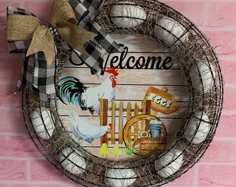 May include: A farmhouse-style wreath with a wire frame, adorned with white eggs and a wooden sign that says "Welcome". The sign features a rooster, hen, and chicks. A buffalo check bow adds a rustic touch.