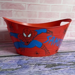 May include: Red plastic storage bin with a Spiderman design. The bin has a handle on each side and features a blue and red Spiderman image with the word "SAM" printed on the side.