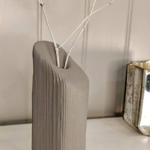 May include: A grey ceramic vase with a textured, wood-like surface. The vase is tall and narrow, with a wide opening at the top. It is filled with white dried branches.