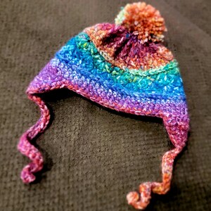 Peut inclure: Un bonnet crocheté fait à la main, coloré, avec des cache-oreilles et un pompon. Le bonnet présente un arc-en-ciel de couleurs vives, notamment le violet, le bleu, le vert et l'orange. Les cache-oreilles ont un design bouclé. Ce bonnet douillet est parfait pour le froid.