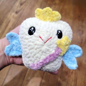 May include: A handmade, plush tooth fairy doll. The doll is white with large black eyes, a red stitched smile, and a yellow crown. It has blue wings and a purple and yellow wand. The doll is made of soft, textured yarn.