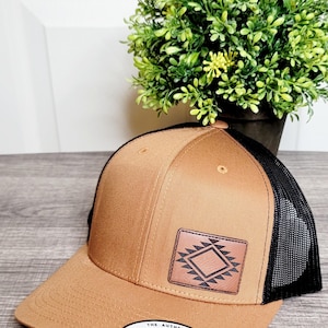 Op de afbeelding: Een bruine en zwarte trucker-pet met een bruine leren patch met een geometrisch ontwerp. De pet heeft een zwart mesh-achterkant en een bruine voorkant met een "The Authentic Yupoong Snapback" sticker aan de onderkant.
