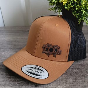 May include: A brown snapback cap with a black mesh back. The cap has a black sunflower design on the front and the text "THE AUTHENTIC SNAPBACK YUPOONG SINCE 1974" on a white oval patch.