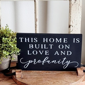 May include: Black wooden sign with white text that reads "This home is built on love and profanity".