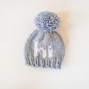 May include: A light blue knitted baby hat with a white pom-pom on top and the letters "HI" knitted in white on the front.