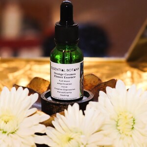 May include: A small green glass bottle with a dropper, labeled "Essential Botany Orange Cosmos Flower Essence". The bottle is sitting on a brown wooden stand with two white flowers on a gold background.