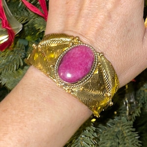 May include: A gold bracelet with a large pink gemstone set in the center. The bracelet is decorated with intricate gold leaf designs.