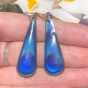 Könnte beinhalten: Ein Paar tropfenförmige Ohrringe mit einem blauen und grünen, irisierenden Finish. Die Ohrringe sind aus Metall und haben eine goldfarbene Oberfläche.