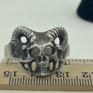 Può includere: Anello d'argento con un designo a forma di teschio e corna di ariete. L'anello è su un righello di legno.