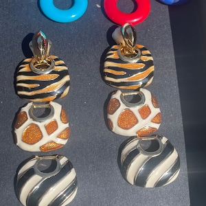 May include: A pair of gold-tone earrings with a layered design. Each earring features three charms: a black and white zebra print, a brown and white cheetah print, and a black and white striped charm.