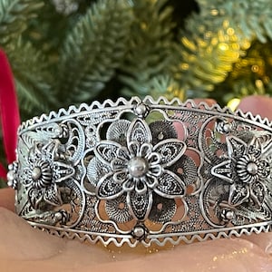 May include: A silver filigree bracelet with a floral design. The bracelet is made of intricate, delicate metalwork and features a series of interconnected flowers.