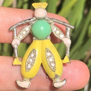 Op de afbeelding: Een gele emaille pin met een grillig ontwerp van een persoon met een groene edelsteen, die een kroon draagt en een stok vasthoudt. De pin is versierd met strass.