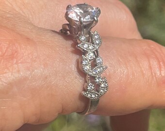 May include: A silver ring with a large, clear, round gemstone surrounded by smaller gemstones. The band is decorated with a leaf design.
