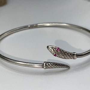 Könnte beinhalten: Ein silbernes Schlangenarmband mit zwei rosafarbenen Rubin-Steinen als Augen.