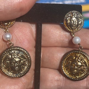 Puede incluir: Pendientes de tono dorado con una gota de perla y un colgante redondo en forma de moneda con un diseño de cabeza de Medusa.