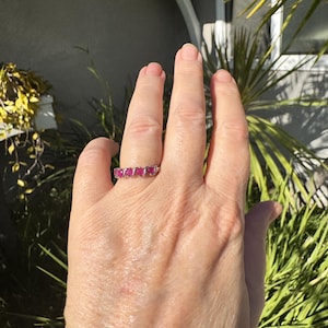 Könnte beinhalten: Ein silberner Ring mit fünf leuchtend roten Edelsteinen in einer Reihe. Der Ring wird auf einer Hand gezeigt, mit einem Hintergrund aus grünem Laub und Sonnenlicht.