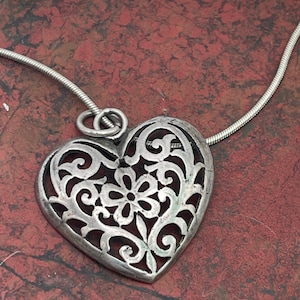 May include: Silver-toned heart pendant necklace with an intricate openwork design. The pendant features a floral pattern at the center and is suspended from a delicate chain. The necklace is displayed on a reddish-brown surface.