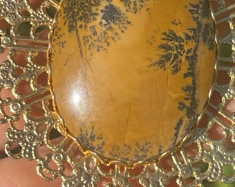 May include: A gold-toned metal pendant with a large oval-shaped stone. The stone is a light brown color with dark brown dendritic patterns.