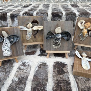 May include: Five decorative wall art pieces featuring angel figures crafted from oyster shells and stones mounted on square or rectangular wooden plaques. Four of the pieces are displayed on small wooden easels, while one lies flat on the surface.