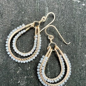 Puede incluir: Un par de pendientes de alambre dorado con cuentas de color azul claro. Los pendientes tienen forma de lágrima y tienen una doble capa de cuentas.