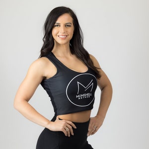Peut inclure: Une femme portant un débardeur noir avec un logo blanc qui dit "Momshell Method". Le logo est à l'intérieur d'un cercle avec un "M" stylisé au centre.