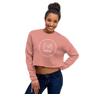 Peut inclure: Un sweat-shirt rose pâle à manches longues et à coupe courte avec un logo circulaire blanc qui dit "Momshell Method" et un "M" stylisé à l'intérieur du cercle.