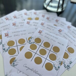 May include: A stack of baby shower scratch-off game cards. Each card features a tic-tac-toe grid with gold circles, the text "SCRATCH TO SEE WHAT Baby House WILL BE," and a "Congratulations!" message. Decorative floral elements are also present.