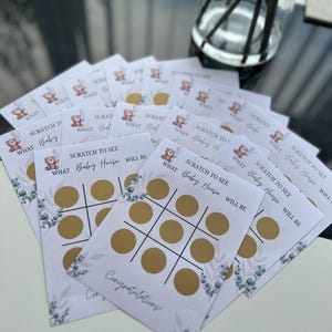May include: A stack of baby shower scratch-off game cards. Each card features a tic-tac-toe grid with gold circles, the text "SCRATCH TO SEE WHAT Baby House WILL BE," and a teddy bear graphic. The cards are white with green floral accents.