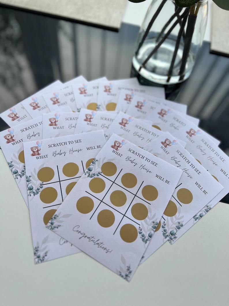 May include: A stack of baby shower scratch-off game cards. Each card features a tic-tac-toe grid with gold circles, the text "SCRATCH TO SEE WHAT Baby House WILL BE," and a teddy bear graphic. The cards are white with green floral accents.