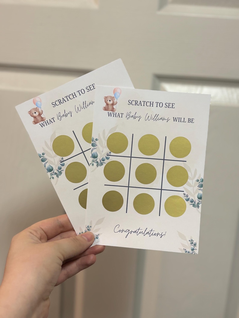May include: Two white scratch-off cards for a baby shower game. Each card has a tic-tac-toe grid with gold circles to scratch. The text "SCRATCH TO SEE WHAT Baby Williams WILL BE" is printed above the grid, with a teddy bear graphic. Floral accents and the word "Congratulations!" are also present.