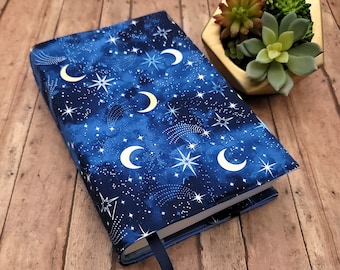 May include: A book cover with a celestial design. The cover features a dark blue background with white crescent moons, stars, and shooting stars. A navy blue ribbon bookmark extends from the book. A small gold geometric planter with succulents is in the background.