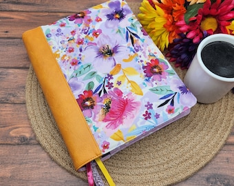 May include: A floral patterned notebook cover with a yellow leather spine and a white background. The cover features a variety of pink, purple, and yellow flowers with green leaves. The notebook is closed and has a yellow ribbon bookmark.