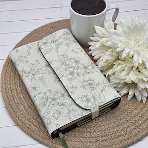 May include: A fabric-covered journal featuring a light cream background with a delicate gray floral line art pattern. The journal has a fold-over flap closure secured by a matching fabric band and includes a thin green ribbon bookmark.
