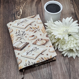 May include: A book cover with a beige background featuring illustrations of open books, spectacles, coffee cups, and autumn leaves. A white mug and white flowers are next to the book cover on a wooden surface.