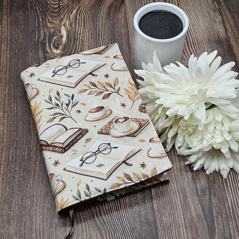 May include: A fabric book cover featuring a repeating pattern of open books, round eyeglasses, coffee cups, and autumn leaves in shades of brown, tan, and cream. The cover is displayed alongside a white ceramic mug and white flowers.