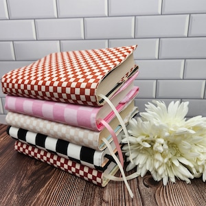 May include: A stack of five books with colorful patterned covers. The top book has a red and white checkered pattern, the second book has a pink and white checkered pattern, the third book has a beige and white checkered pattern, the fourth book has a black and white checkered pattern, and the fifth book has a red and white checkered pattern.