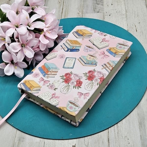 Könnte beinhalten: Ein Hardcover-Buch mit rosa Einband, verziert mit Illustrationen von Büchern, Teetassen und Blumenarrangements. Ein rosa Lesezeichen aus Band ragt aus dem Buch. Das Buch liegt auf einer türkisfarbenen Oberfläche mit rosa Blumen im Hintergrund.