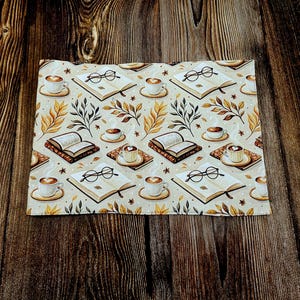 May include: A rectangular fabric placemat featuring a repeating pattern of open books, pairs of black-rimmed reading glasses, and steaming cups of coffee on saucers, interspersed with small brown and yellow autumn leaves on a cream background.