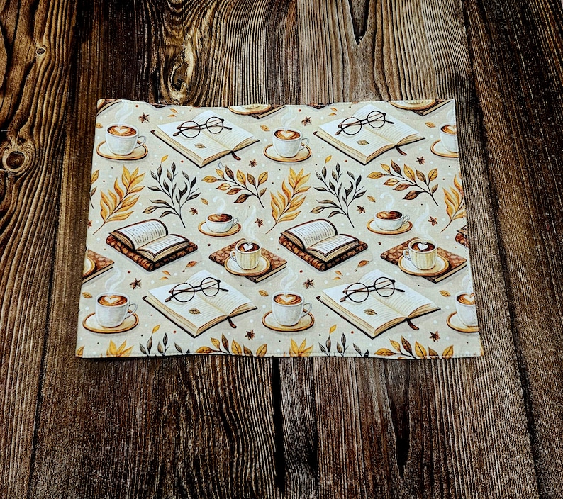 May include: A rectangular fabric placemat featuring a repeating pattern of open books, pairs of black-rimmed reading glasses, and steaming cups of coffee on saucers, interspersed with small brown and yellow autumn leaves on a cream background.