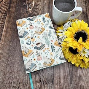 May include: A patterned journal with a teal ribbon, featuring a repeating design of cats, books, coffee cups, and floral elements. A white mug with dark liquid, and a bouquet of sunflowers and daisies are also present.