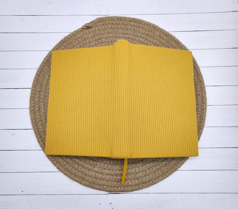 May include: A yellow ribbed notebook with a yellow ribbon bookmark. The notebook is sitting on a woven straw mat.