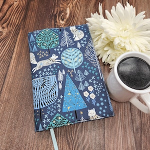 May include: A rectangular fabric book cover featuring a dark blue background with a repeating pattern of white foxes, stylized light blue and teal trees, and small geometric shapes. A light blue ribbon bookmark extends from the bottom edge.