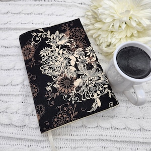 May include: A black fabric journal cover with a cream and brown floral pattern. The cover has a cream colored spine and a white ribbon bookmark.