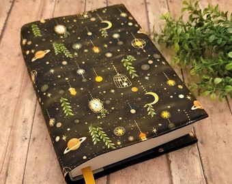 May include: A black book cover with a celestial pattern featuring planets, moons, stars, and hanging lanterns. The cover has a yellow bookmark and is on a wooden surface. The design includes gold, white, and green accents.