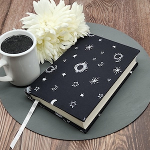 May include: A black journal with a celestial design featuring white stars, moons, and suns. A white ribbon bookmark extends from the book. A white mug filled with dark liquid and a white flower are also present.